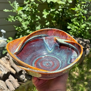 May include: A ceramic bowl featuring a deep, glossy glaze with swirling patterns of dark red, burnt orange, and teal blue. The bowl has a wide, open rim and a curved, integrated handle that extends across the top edge, creating a unique silhouette.