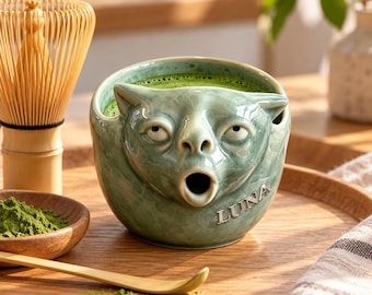 Może przedstawiać: Jasnozielony ceramiczny kubek w kształcie kociego pyszczka z wytłoczonym napisem LUNA z boku. Kubek jest wypełniony zieloną herbatą matcha i stoi obok bambusowej trzepaczki, małej drewnianej miseczki z proszkiem matcha oraz drewnianej łyżeczki.