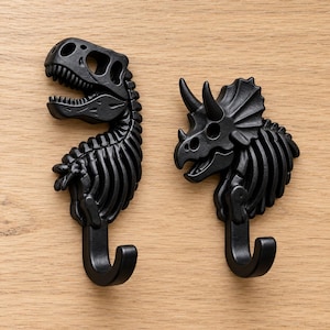 May include: Two matte black wall hooks shaped like dinosaur skeletons. The left hook features a T-Rex skull and ribcage, while the right hook depicts a Triceratops skull and ribcage. Each piece includes a curved hook at the base.
