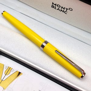 Op de afbeelding: Een felgele vulpen met een gepolijste zilverkleurige clip en middenring, rustend in een witte presentatiedoos met de merknaam Montblanc in zwarte letters gedrukt op de voering van het binnendeksel.