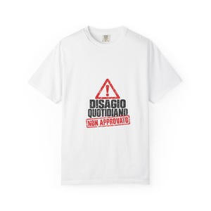 Può includere: Una t-shirt bianca a maniche corte con un segnale di pericolo triangolare rosso con un punto esclamativo. Sotto il segnale, il testo recita DISAGIO QUOTIDIANO in nero, seguito da un timbro rettangolare rosso NON APPROVATO.