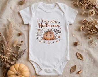 Puede incluir: Un body blanco de manga corta para bebé con un estampado gráfico que incluye el texto "Il mio primo Halloween" sobre una calabaza sonriente, un fantasma, un murciélago, una araña y hojas de otoño en tonos naranja, marrón y gris.