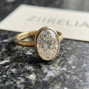 Puede incluir: Un anillo de oro con una gran piedra preciosa transparente de talla ovalada y facetada, montada en un engaste de bisel liso. La banda es de oro pulido. En el fondo, una tarjeta blanca muestra el texto ZIRELIA en letras mayúsculas.