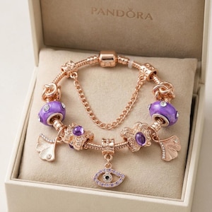Può includere: Un bracciale con ciondoli in oro rosa caratterizzato da perline in vetro viola, ciondoli metallici a forma di foglia di ginkgo e un pendente centrale a forma di occhio con piccole pietre chiare, presentato in una scatola beige Pandora.