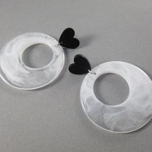 May include: A pair of large, clear acrylic earrings with a white, marbled effect. Each earring has a black heart-shaped accent at the top. The earrings have a circular design with a hole in the centre, creating a modern, minimalist aesthetic.