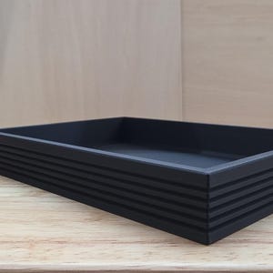 May include: A rectangular black tray with a matte finish and a series of horizontal grooves along the exterior side walls. The tray features a flat base and raised edges, providing a structured and open container design for various items.