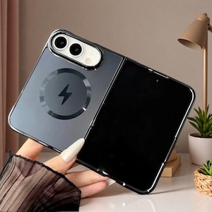 Peut inclure: Une coque de téléphone gris foncé semi-transparente pour smartphone pliable. La coque présente un anneau de charge magnétique circulaire avec une icône d'éclair au centre, une découpe pour deux objectifs et une finition brillante.
