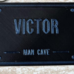 Op de afbeelding: Een rechthoekig zwart bord met een blauwe rand en vier blauwe accenten in de hoeken. De tekst VICTOR is in reli&euml;f aangebracht in grote blauwe letters, met daaronder de tekst MAN CAVE in kleinere blauwe letters.