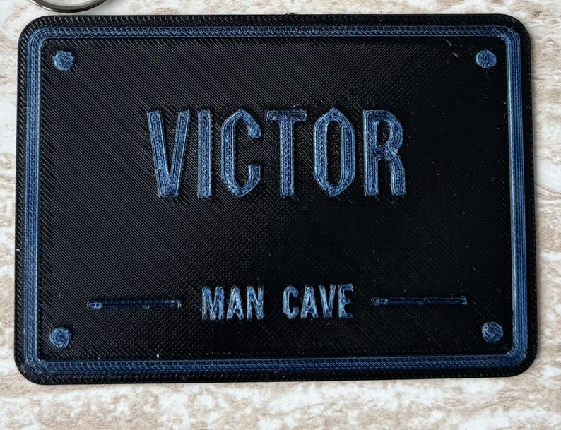 Op de afbeelding: Een rechthoekig zwart bord met een blauwe rand en vier blauwe accenten in de hoeken. De tekst VICTOR is in reli&euml;f aangebracht in grote blauwe letters, met daaronder de tekst MAN CAVE in kleinere blauwe letters.