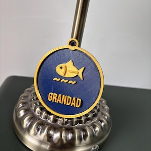 May include: A circular navy blue pendant with a yellow border and a small loop at the top. The center features a yellow fish icon above three yellow wavy lines and the word GRANDAD in raised yellow capital letters.