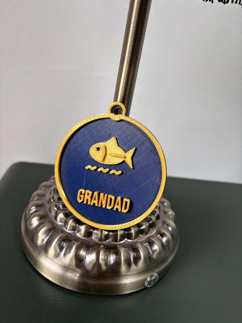 May include: A circular navy blue pendant with a yellow border and a small loop at the top. The center features a yellow fish icon above three yellow wavy lines and the word GRANDAD in raised yellow capital letters.