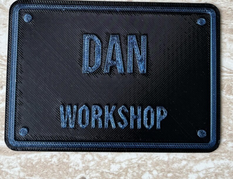 Op de afbeelding: Een rechthoekig zwart 3D-geprint bord met een blauwe rand en vier ronde accenten in de hoeken. De tekst DAN is in het midden in reli&euml;f aangebracht, met het woord WORKSHOP er direct onder in een bijpassend blauw schreefloos lettertype.