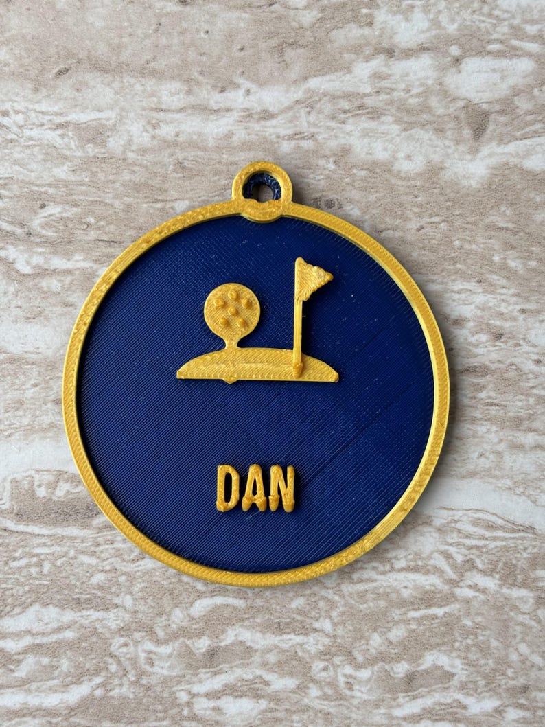 Puede incluir: Un adorno circular con un centro azul oscuro y un borde dorado. El centro presenta un gr&aacute;fico dorado impreso en 3D de una pelota de golf sobre un tee junto a una bandera en un green. El nombre DAN est&aacute; impreso en letras doradas.