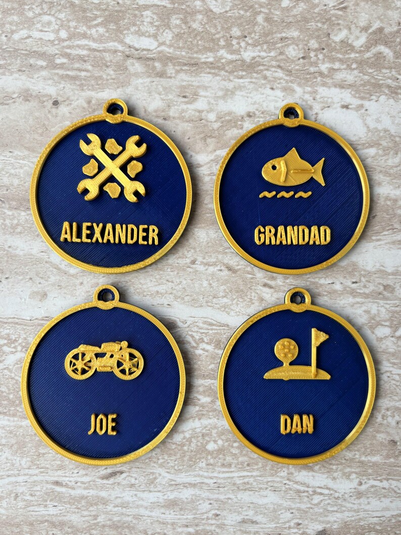 May include: Four circular ornaments with a dark blue center and gold rim. Each features a gold icon and name: Alexander with crossed wrenches, Grandad with a fish, Joe with a motorcycle, and Dan with a golf ball and flag. Each has a small loop at the top.