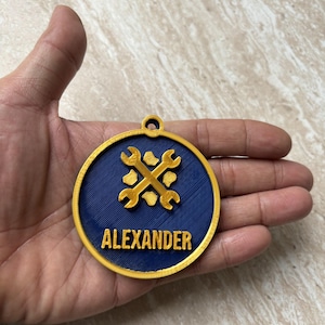 May include: A circular gold and dark blue medallion featuring a raised gold wrench icon and the name ALEXANDER in gold lettering. The medallion has a small loop at the top for hanging and a textured finish on the blue surface.