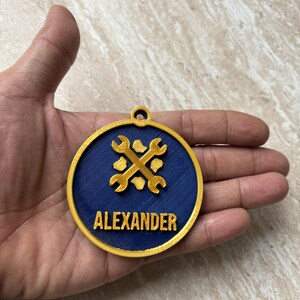 Puede incluir: Un medall&oacute;n circular dorado y azul oscuro con un icono de llave inglesa dorada en relieve y el nombre ALEXANDER en letras doradas. El medall&oacute;n tiene una peque&ntilde;a anilla en la parte superior para colgarlo y una superficie azul texturizada.