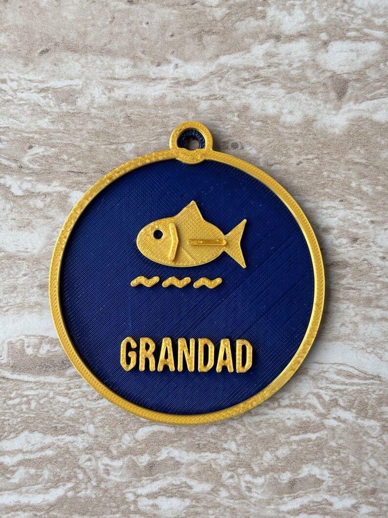 May include: A circular ornament with a dark blue center and a gold border. It features a gold fish icon above three gold wavy lines and the word GRANDAD in gold capital letters. A small gold loop is at the top for hanging purposes.