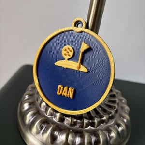 Puede incluir: Un medall&oacute;n circular impreso en 3D con fondo azul oscuro, borde dorado y un relieve en color oro que muestra una pelota de golf, una bandera y el nombre DAN. El medall&oacute;n incluye una peque&ntilde;a anilla en la parte superior para colgarlo.