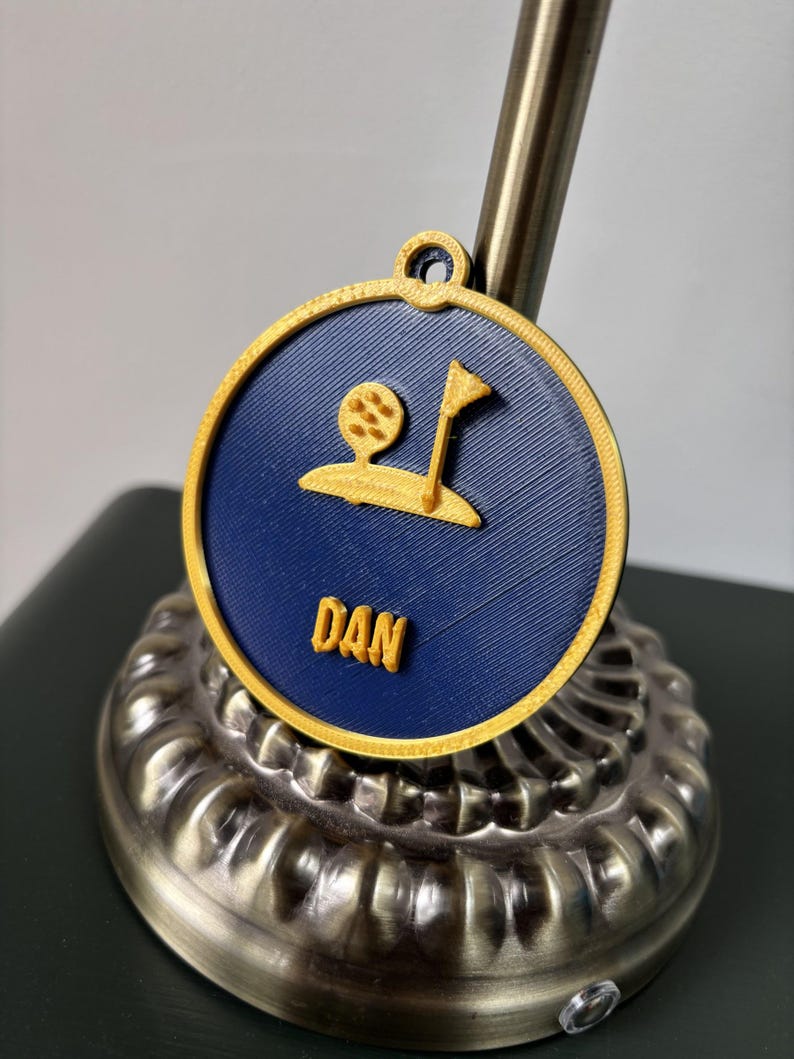 Puede incluir: Un medall&oacute;n circular impreso en 3D con fondo azul oscuro, borde dorado y un relieve en color oro que muestra una pelota de golf, una bandera y el nombre DAN. El medall&oacute;n incluye una peque&ntilde;a anilla en la parte superior para colgarlo.