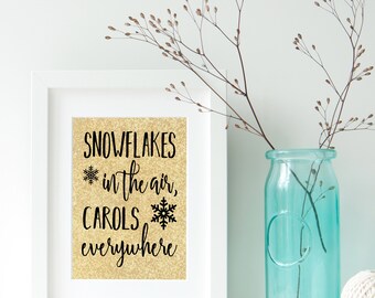 May include: A white framed print with a gold glitter background and black text that reads "Snowflakes in the air, carols everywhere". The print is sitting on a white surface with a blue glass vase and a white ball of twine.