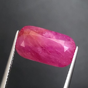 May include: A vibrant, rectangular-cut pink gemstone with a faceted surface, held securely by a pair of silver-toned metal tweezers. The stone displays a deep, saturated magenta hue with internal inclusions and light-reflecting facets.