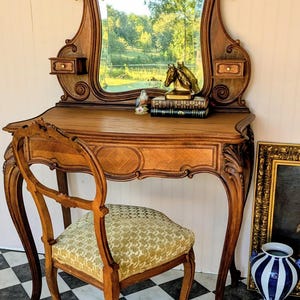 May include: A wooden vanity table with a large arched mirror, two small side drawers, and curved legs. A matching wooden chair with a gold patterned upholstered seat is placed in front. The vanity surface holds a small bird figurine and a brass horse head sculpture resting on two books.