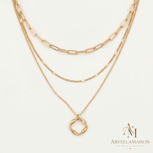 Puede incluir: Un collar de capas en tono dorado que presenta tres cadenas distintas: una cadena de eslabones rectangulares, una cadena texturizada y una cadena fina con un colgante cuadrado retorcido. El logotipo ArvellaMaison es visible.