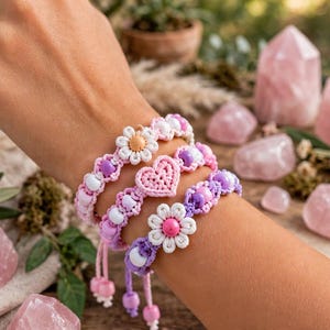 Könnte beinhalten: Drei handgefertigte Makramee-Armbänder mit pastellfarbenen violetten, rosa und weißen Perlen. Ein Armband hat ein zentrales gehäkeltes Herz, während die anderen beiden weiße gänseblümchenförmige Anhänger mit rosa bzw. gelben Zentren aufweisen.