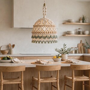 Puede incluir: Una lámpara colgante de macramé con una pantalla en forma de cúpula hecha de cuerda color crema, decorada con cuentas de madera natural y un borde de flecos verde salvia, suspendida por un cordón trenzado con un soporte de latón.