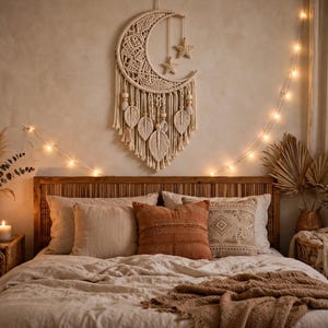May include: A cream-colored macrame wall hanging shaped like a crescent moon with two hanging stars, leaf-shaped fringe, and wooden beads. The piece is suspended against a wall above a bed with neutral-toned pillows and a textured throw blanket.
