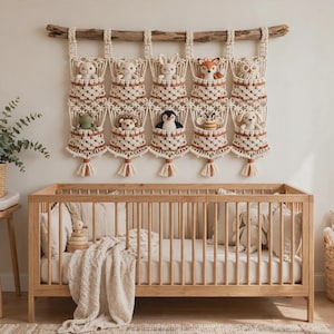 Op de afbeelding: Een macramé wandhanger met tien individuele vakjes die aan een houten tak hangen. Elk vakje bevat een kleine pluche dierenknuffel. De organizer is gemaakt van gebroken wit koord met bruine accenten en heeft kwastjes aan de onderkant.