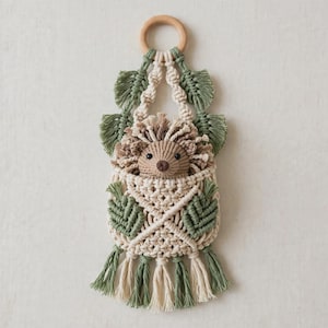 May include: A macrame wall hanging featuring a central hedgehog figure made of cream and brown cord. The design includes sage green leaf-shaped accents, a circular wooden ring at the top for hanging, and a decorative fringe at the bottom.