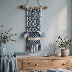 Op de afbeelding: Een macramé wandhanger met een blauw en crème geknoopt patroon, opgehangen aan een stuk drijfhout. Het ontwerp bevat een centraal vak, twee zijflappen en een onderste franje gemaakt van gedraaid katoenen koord in blauw en crème.