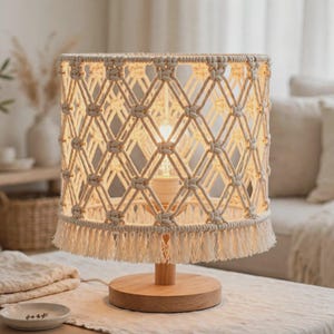 May include: A table lamp featuring a cylindrical shade made of off-white macrame cord with a diamond knot pattern and a fringed bottom edge. The lamp has a light-colored wooden base and a central stem holding a single glowing light bulb.