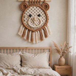 May include: A macrame wall hanging shaped like a bear face, crafted from cream and brown cotton cord. It features two circular ears, dark round eyes, a central wooden bead nose, and a fringe of alternating cream and brown tassels.