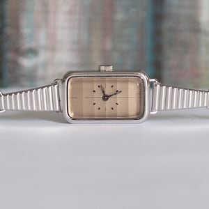 May include: A silver watch with a square face and a brown dial. The watch has a silver metal band.