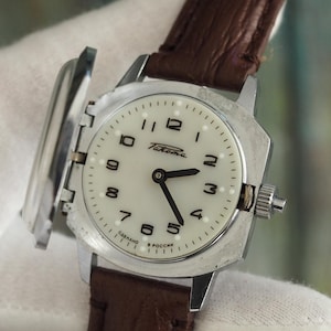 Könnte beinhalten: Eine Vintage-Armbanduhr mit einem quadratischen, silberfarbenen Gehäuse und einem weißen Zifferblatt mit schwarzen Ziffern und Zeigern. Die Uhr hat ein braunes Lederarmband. Die Uhr wird von einer behandschuhten Hand gehalten.