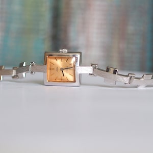 Puede incluir: Un reloj plateado con una esfera cuadrada y una correa plateada. La esfera del reloj es de color dorado claro con agujas y marcadores negros.