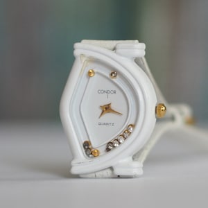 May include: White analog wristwatch with a gold Condor logo and gold accents. The watch face is white with gold hands and a white quartz inscription.