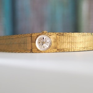 May include: A gold-toned wristwatch with a round face and a gold metal band. The band is a woven design.