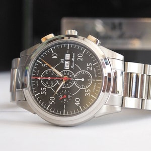 May include: A silver chronograph wristwatch with a black face and red accents. The watch has a date window and a tachymeter scale. The watch is on a silver metal bracelet.