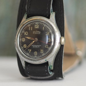 Puede incluir: Un reloj de pulsera plateado con una esfera negra y una correa de cuero negra. La esfera del reloj tiene números y manecillas blancas, y las palabras "DELGAMA" y "17 JEWELS" están impresas en ella.