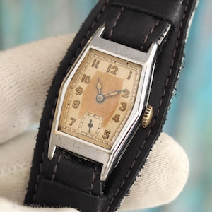 May include: A vintage wristwatch with a silver rectangular case and a light brown face. The watch has black numerals and hands, set against a light brown background. It is mounted on a black leather cuff-style strap.