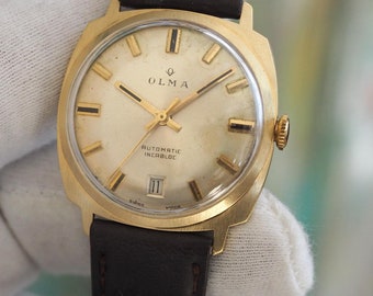 May include: A gold-toned wristwatch with a square face and a brown leather band. The watch face has a white background with black Roman numeral hour markers and black hands. The watch face also has the text "Olma Automatic Incabloc" printed on it.