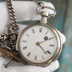 Könnte beinhalten: Eine silberne Taschenuhr mit weißem Zifferblatt und schwarzen römischen Ziffern. Die Uhr hat eine Kette und einen kleinen goldfarbenen Gewehranhänger. Die Uhr wird von einer behandschuhten Hand gehalten.