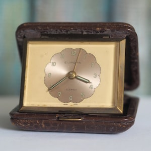 Op de afbeelding: Een vintage bruine leren reiswekker met een gouden wijzerplaat en het woord "EUROPA" gedrukt op de wijzerplaat.