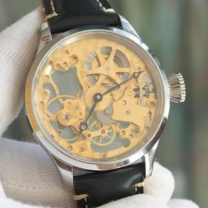 May include: A silver-toned skeleton watch with a gold-colored face, showcasing the intricate inner workings. The watch features a black leather band with white stitching. The watch is a classic timepiece.