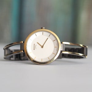 Puede incluir: Un reloj de pulsera plateado y dorado con una esfera blanca y agujas negras. El reloj tiene el logotipo de Boccia en la esfera.