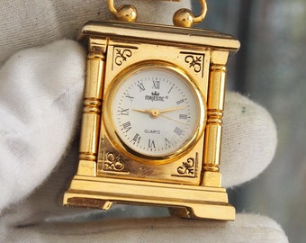 May include: Gold-toned tabletop clock with a white face and Roman numeral hour markers. The clock face says "Majestic" and "Quartz".