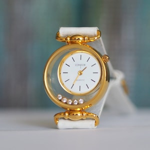 May include: A gold-tone Condor quartz wristwatch with a white face and a white leather band. The watch has a clear case with a row of small rhinestones around the edge.